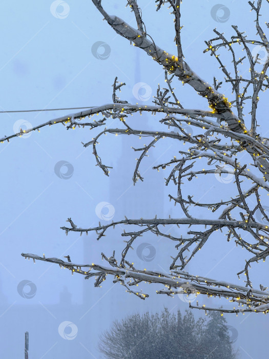 Скачать Московский государственный университет имени М.В. Ломоносова виден сквозь густой зимний туман во время снегопада. На переднем плане стоит украшенная елка со светящимися гирляндами фотосток Ozero