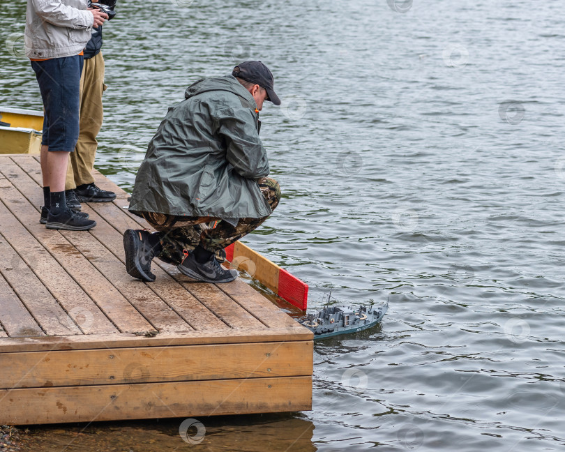 Скачать Россия,город Железногорск Красноярский Край, 26 августа 2023 год. Мужчина сидит на корточках у воды. В воде — небольшая модель корабля. фотосток Ozero