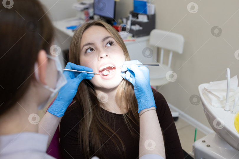 Скачать Симпатичная молодая женщина-стоматолог проводит обследование и лечение молодой пациентки в стоматологической клинике. фотосток Ozero