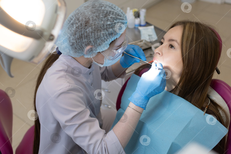 Скачать Симпатичная молодая женщина-стоматолог проводит обследование и лечение молодой пациентки в стоматологической клинике. фотосток Ozero