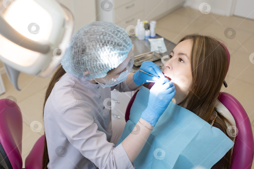 Скачать Симпатичная молодая женщина-стоматолог проводит обследование и лечение молодой пациентки в стоматологической клинике. фотосток Ozero