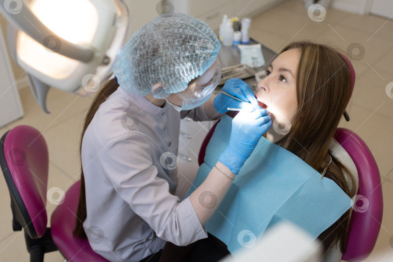 Скачать Симпатичная молодая женщина-стоматолог проводит обследование и лечение молодой пациентки в стоматологической клинике. фотосток Ozero