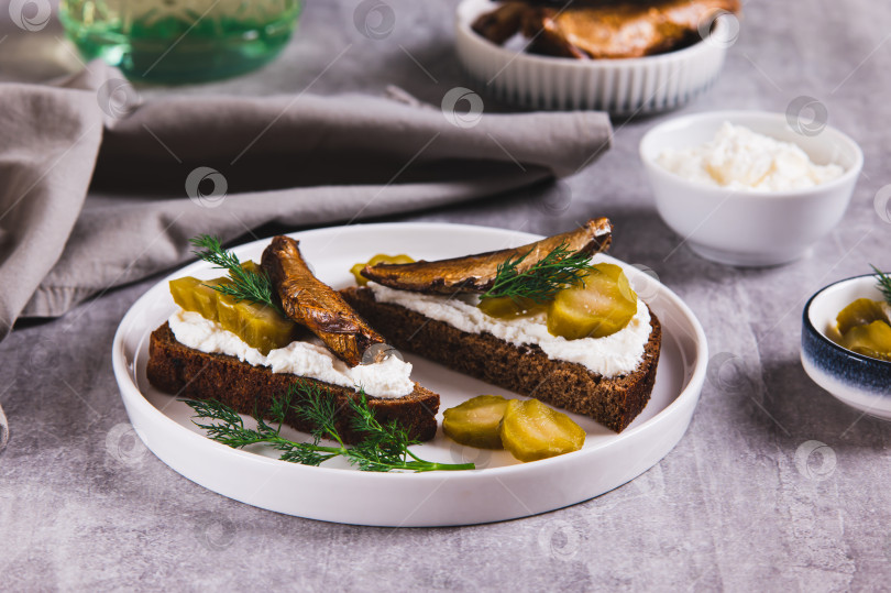 Скачать Бутерброды с рикоттой, солеными огурцами и шпротами на ржаном хлебе на тарелке на столе фотосток Ozero