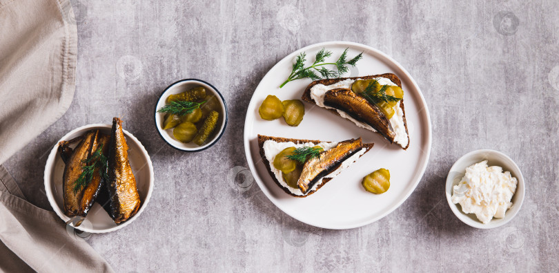 Скачать Тосты из ржаного хлеба со сливочным сыром, шпротами и солеными огурцами на тарелке на столешнице посмотреть веб-баннер фотосток Ozero