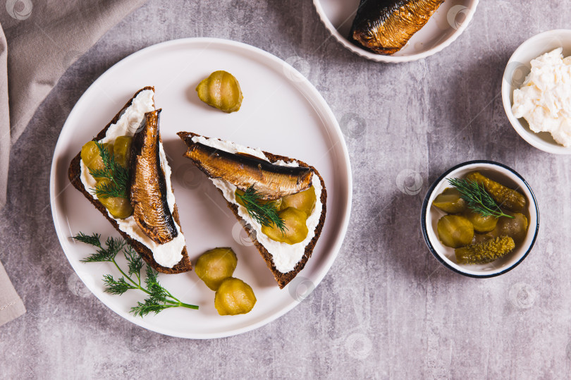 Скачать Бутерброды с рикоттой, солеными огурцами и шпротами на ржаном хлебе на тарелке на столе вид сверху фотосток Ozero