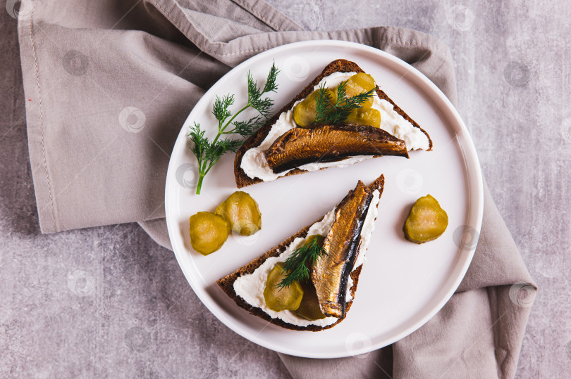 Скачать Бутерброды с рикоттой, солеными огурцами и шпротами на ржаном хлебе крупным планом на тарелке на столе, вид сверху фотосток Ozero