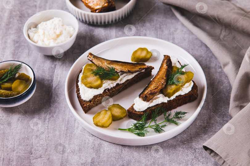 Скачать Тосты из ржаного хлеба со сливочным сыром, шпротами и солеными огурцами на тарелке на столе фотосток Ozero