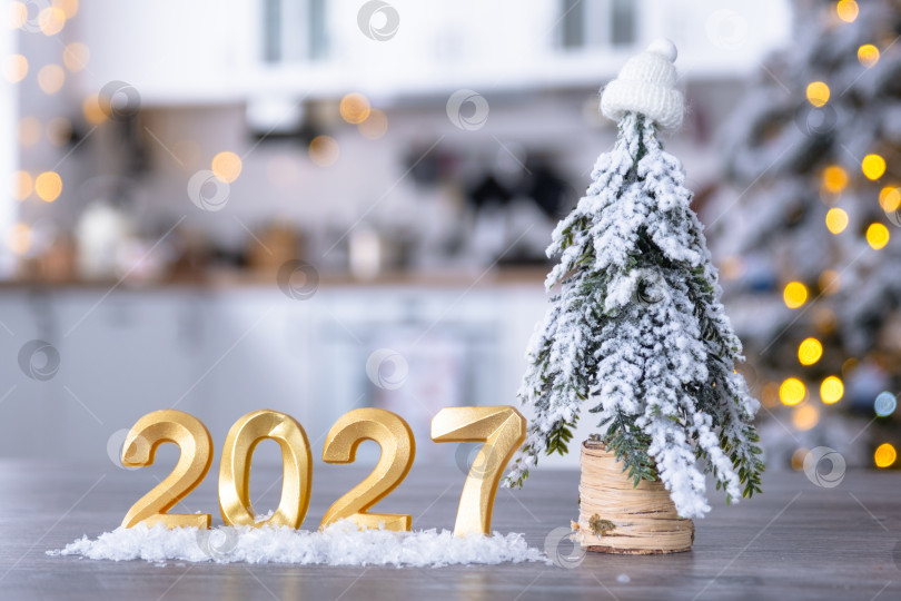 Скачать Золотая цифра 2027 на столе на фоне уютной белой кухни с рождественской елкой и рождественским декором из гирлянд. Жилье, интерьер, ремонт, ипотека, аренда в Новом году фотосток Ozero