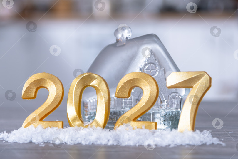 Скачать Золотые фигурки с цифрами Нового 2027 года на фоне крошечного домика с теплым светом и гирляндами. Поздравительная открытка, С Новым годом, уютный дом фотосток Ozero