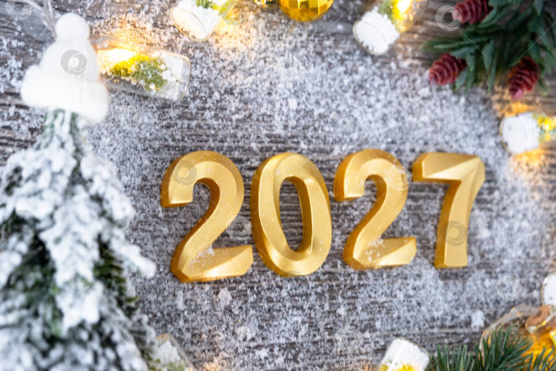 Скачать Поздравительные золотые цифры с Новым 2027 годом на уютном, праздничном сером деревянном фоне с рождественской елкой, снегом, огнями гирлянд. Поздравления, открытка. Календарь, обложка фотосток Ozero