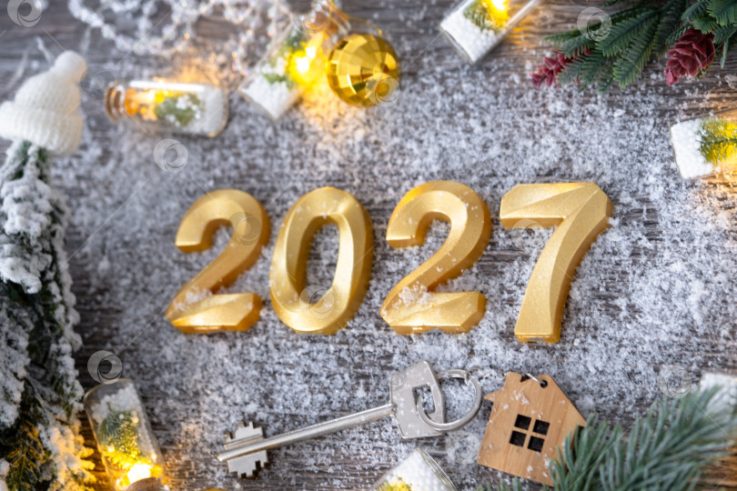 Скачать Золотые цифры на Новый 2027 год и ключ от дома с миниатюрной формой дома на праздничном сером деревянном фоне, флэтлэй, гирлянды, поздравительная открытка. Жилье, недвижимость, переезд, ипотека, страхование. фотосток Ozero
