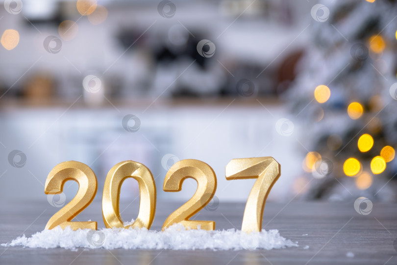Скачать Золотая цифра 2027 на столе на фоне уютной белой кухни с рождественской елкой и рождественским декором из гирлянд. Жилье, интерьер, ремонт, ипотека, аренда в Новом году фотосток Ozero