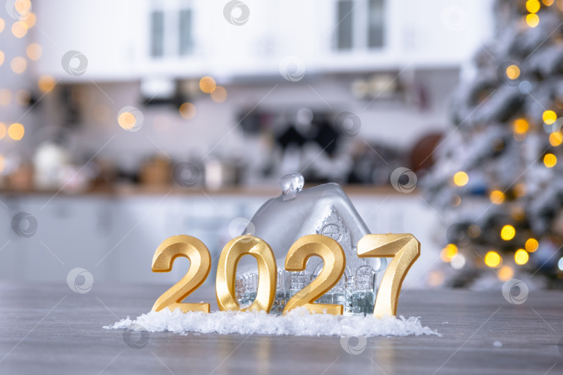 Скачать Золотая цифра 2027 и крошечный домик на фоне уютной белой кухни с рождественской елкой и рождественским декором из гирлянд. Жилье, интерьер, ремонт, ипотека, аренда в Новом году фотосток Ozero