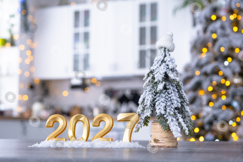Скачать Золотая цифра 2027 на столе на фоне уютной белой кухни с рождественской елкой и рождественским декором из гирлянд. Жилье, интерьер, ремонт, ипотека, аренда в Новом году фотосток Ozero