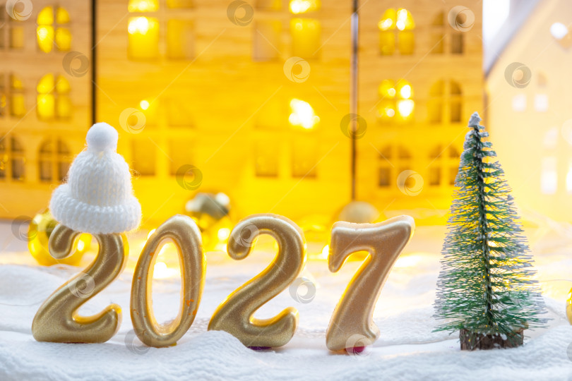 Скачать Золотые цифры, обозначающие Новый 2027 год, на фоне крошечного домика с теплым светом и гирляндами. Поздравительная открытка, С Новым годом, уютный дом фотосток Ozero