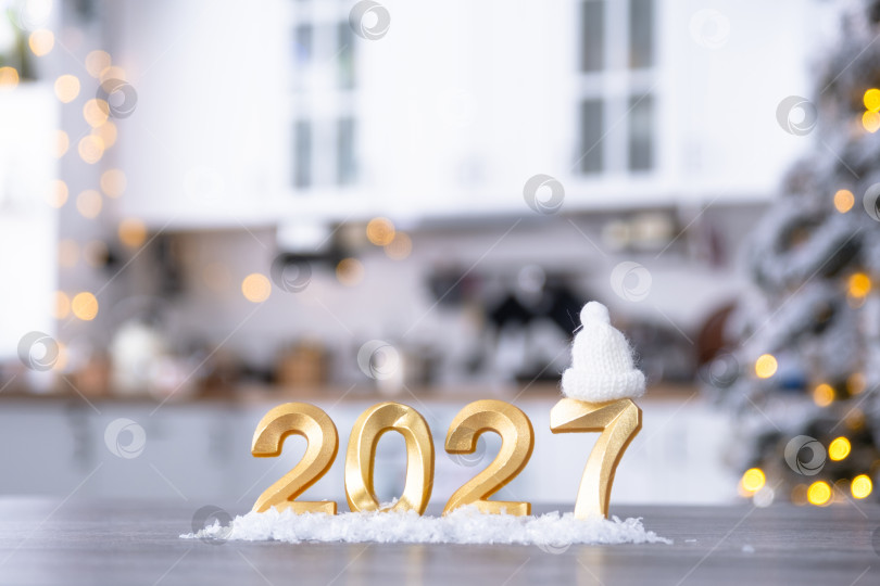 Скачать Золотая цифра 2027 на столе на фоне уютной белой кухни с рождественской елкой и рождественским декором из гирлянд. Жилье, интерьер, ремонт, ипотека, аренда в Новом году фотосток Ozero