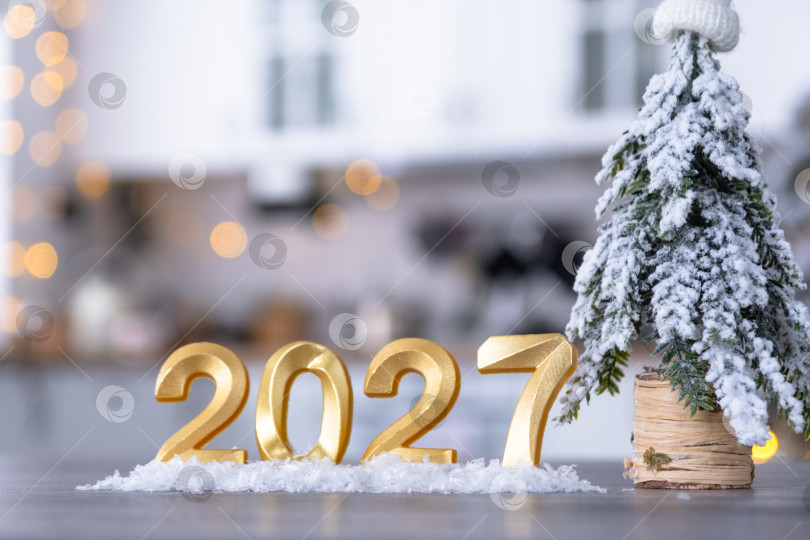 Скачать Золотая цифра 2027 на столе на фоне уютной белой кухни с рождественской елкой и рождественским декором из гирлянд. Жилье, интерьер, ремонт, ипотека, аренда в Новом году фотосток Ozero