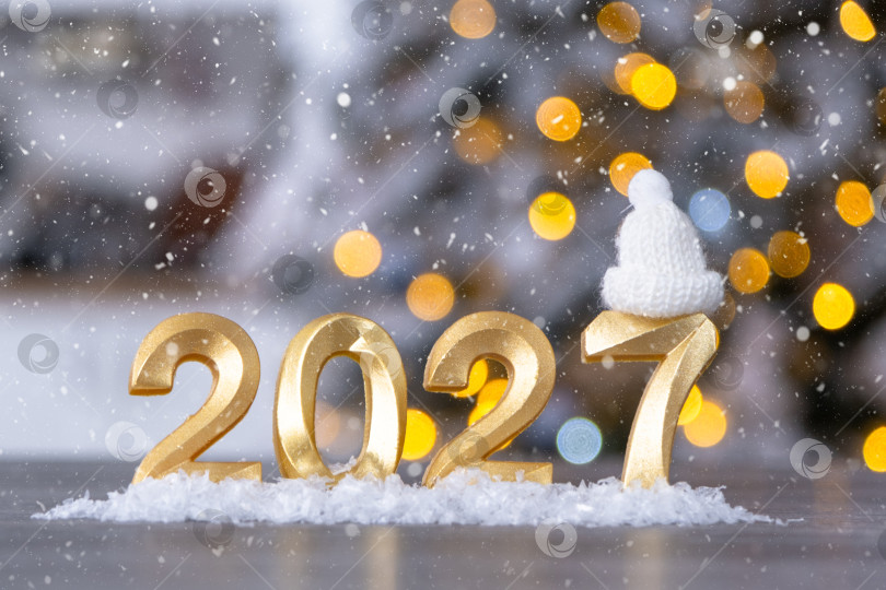 Скачать Золотые фигурки с цифрами Нового 2027 года на фоне сказочного света, снега и гирлянд. Поздравительная открытка, С Новым годом, уютный дом фотосток Ozero