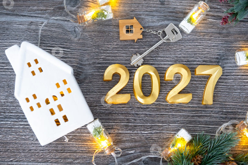 Скачать Золотые цифры на Новый 2027 год и ключ от дома с миниатюрной формой дома на праздничном сером деревянном фоне, флэтлей, гирлянды, поздравительная открытка. Жилье, недвижимость, переезд, ипотека, страхование. фотосток Ozero