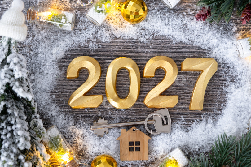 Скачать Золотые цифры на Новый 2027 год и ключ от дома с миниатюрной формой дома на праздничном сером деревянном фоне, флэтлэй, гирлянды, поздравительная открытка. Жилье, недвижимость, переезд, ипотека, страхование. фотосток Ozero