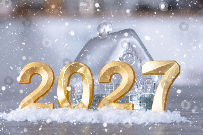 Скачать Золотые фигурки с цифрами Нового 2027 года на фоне крошечного домика с теплым светом и гирляндами. Поздравительная открытка, С Новым годом, уютный дом фотосток Ozero