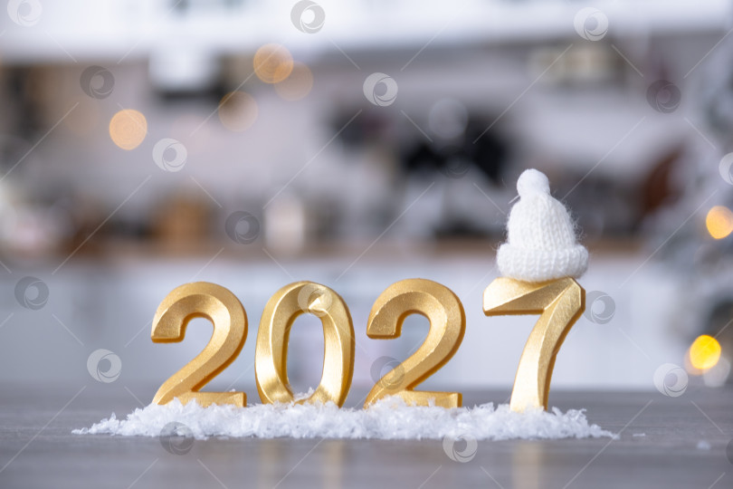 Скачать Золотая цифра 2027 на столе на фоне уютной белой кухни с рождественской елкой и рождественским декором из гирлянд. Жилье, интерьер, ремонт, ипотека, аренда в Новом году фотосток Ozero