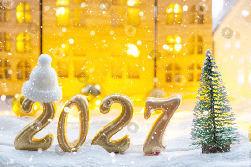 Скачать Золотые фигурки с цифрами Нового 2027 года на фоне крошечного домика с теплым светом и гирляндами. Поздравительная открытка, С Новым годом, уютный дом фотосток Ozero