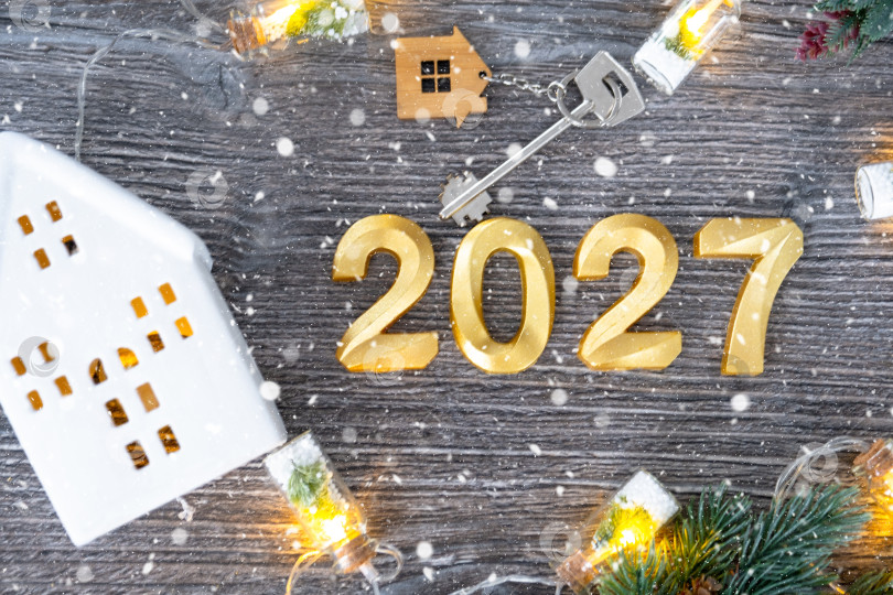 Скачать Золотые цифры на Новый 2027 год и ключ от дома с миниатюрной формой дома на праздничном сером деревянном фоне, флэтлэй, гирлянды, поздравительная открытка. Жилье, недвижимость, переезд, ипотека, страхование. фотосток Ozero