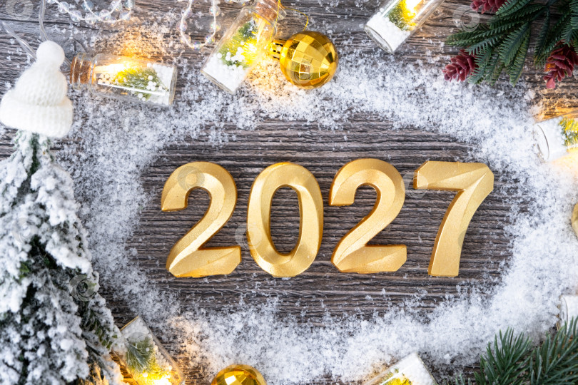 Скачать Поздравительные золотые цифры с Новым 2027 годом на уютном, праздничном сером деревянном фоне с рождественской елкой, снегом, огнями гирлянд. Поздравления, открытка. Календарь, обложка фотосток Ozero