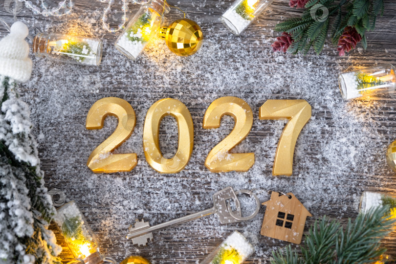 Скачать Золотые цифры на Новый 2027 год и ключ от дома с миниатюрной формой дома на праздничном сером деревянном фоне, флэтлэй, гирлянды, поздравительная открытка. Жилье, недвижимость, переезд, ипотека, страхование. фотосток Ozero