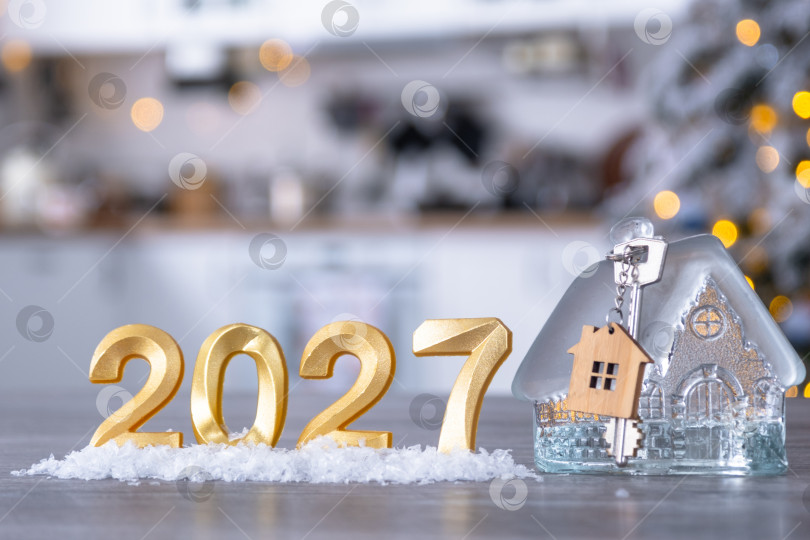 Скачать Золотое число 2027, ключ и крошечный домик на фоне уютной белой кухни с рождественской елкой и рождественским декором из гирлянд. Жилье, интерьер, ремонт, ипотека, аренда в Новом году фотосток Ozero