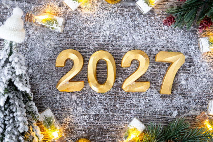 Скачать Поздравительные золотые цифры с Новым 2027 годом на уютном, праздничном сером деревянном фоне с рождественской елкой, снегом, огнями гирлянд. Поздравления, открытка. Календарь, обложка фотосток Ozero