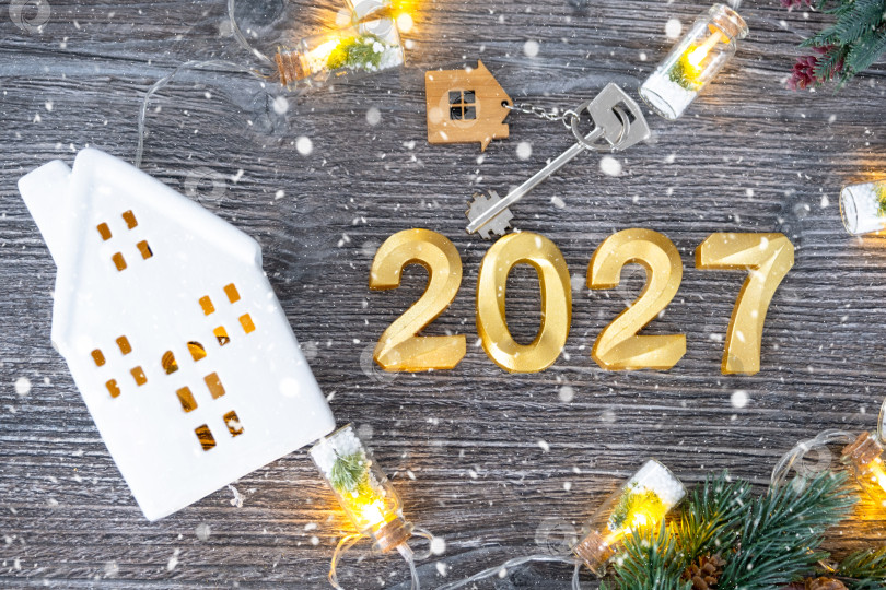 Скачать Золотые цифры на Новый 2027 год и ключ от дома с миниатюрной формой дома на праздничном сером деревянном фоне, флэтлэй, гирлянды, поздравительная открытка. Жилье, недвижимость, переезд, ипотека, страхование. фотосток Ozero