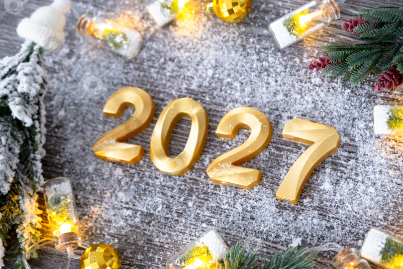 Скачать Поздравительные золотые цифры с Новым 2027 годом на уютном, праздничном сером деревянном фоне с рождественской елкой, снегом, огнями гирлянд. Поздравления, открытка. Календарь, обложка фотосток Ozero