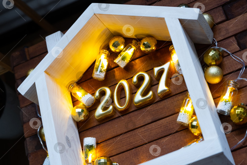Скачать Золотые цифры числа 2027 и крошечный домик на фоне уютных окон дома, залитых теплым светом, с праздничным декором из звезд,снега и гирлянд. Поздравительная открытка, С Новым годом, уютный дом фотосток Ozero