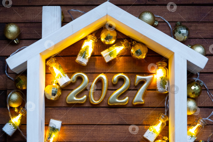 Скачать Золотые цифры числа 2027 и крошечный домик на фоне уютных окон дома, залитых теплым светом, с праздничным декором из звезд,снега и гирлянд. Поздравительная открытка, С Новым годом, уютный дом фотосток Ozero