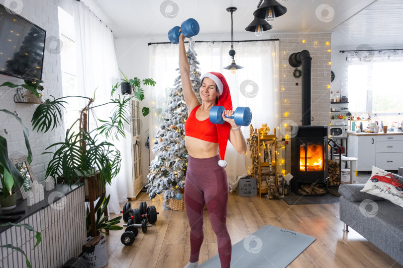 Скачать Женщина в спортивной форме и шапке Санты занимается фитнесом и силовыми упражнениями с гантелями дома в рождественском интерьере с рождественской елкой. Восстановление спортивной формы после Нового года фотосток Ozero
