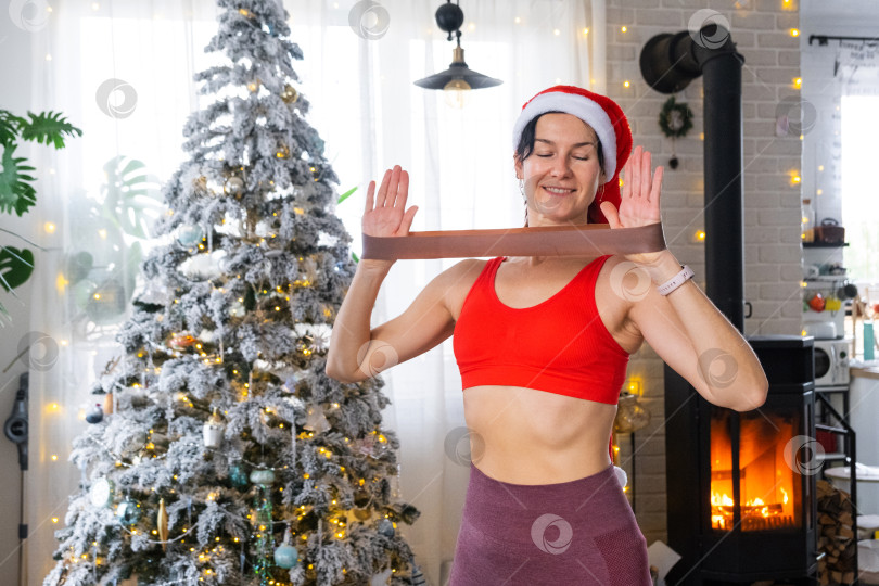 Скачать Женщина в спортивной форме и шапке Санты занимается фитнесом и силовыми упражнениями с фитнес-резинками дома в рождественском интерьере с рождественской елкой. Восстановление спортивной формы после Нового года фотосток Ozero