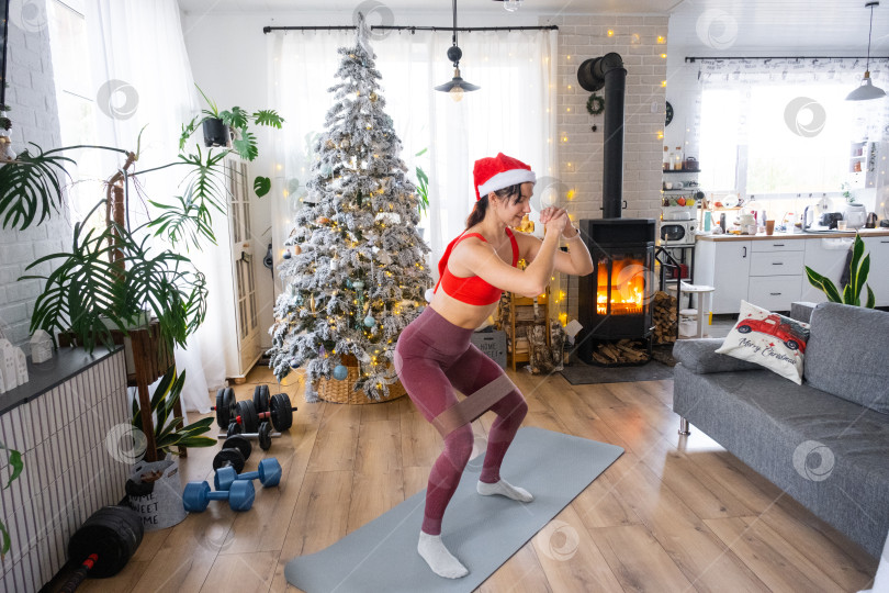 Скачать Женщина в спортивной форме и шапке Санты занимается фитнесом и силовыми упражнениями с фитнес-резинками дома в рождественском интерьере с рождественской елкой. Восстановление спортивной формы после Нового года фотосток Ozero