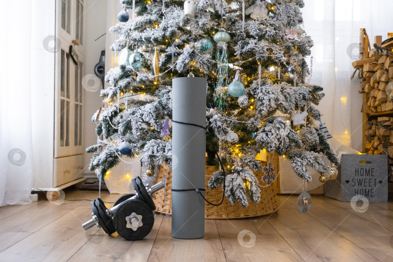 Скачать Спортивный инвентарь - гантели, коврик для фитнеса и ленты для фитнеса - лежат дома под рождественской елкой в качестве подарка на Рождество и Новый год фотосток Ozero