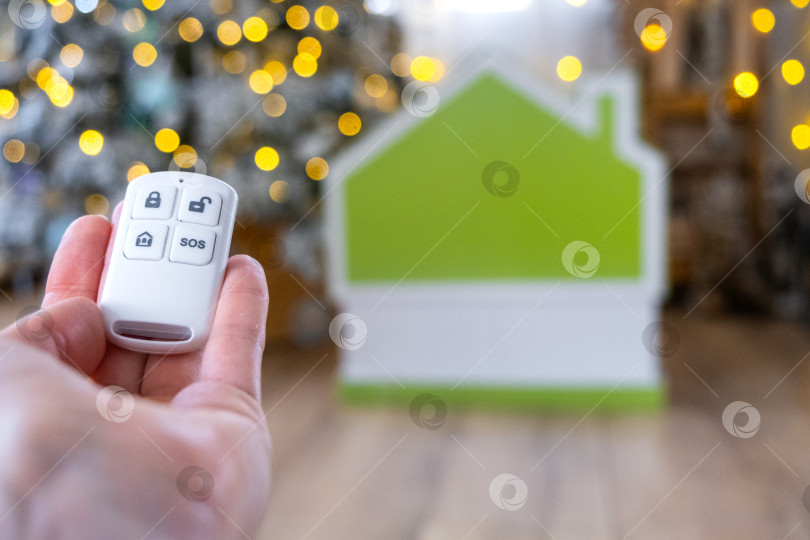 Скачать Брелок для системы безопасности в руке на фоне гирлянды рождественской елки и крошечного домика - подарок на Новый год, Рождество. Безопасность дома, сигнализация, сигнал SOS, защита от воров фотосток Ozero