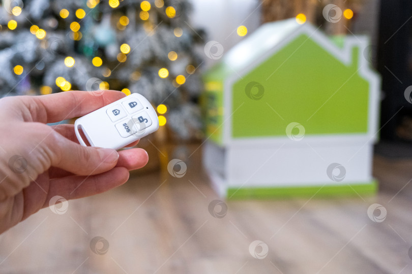 Скачать Брелок для системы безопасности в руке на фоне гирлянды рождественской елки и крошечного домика - подарок на Новый год, Рождество. Безопасность дома, сигнализация, сигнал SOS, защита от воров фотосток Ozero