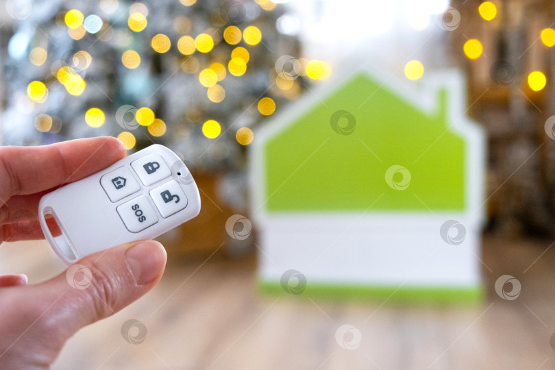 Скачать Брелок для системы безопасности в руке на фоне гирлянды рождественской елки и крошечного домика - подарок на Новый год, Рождество. Безопасность дома, сигнализация, сигнал SOS, защита от воров фотосток Ozero