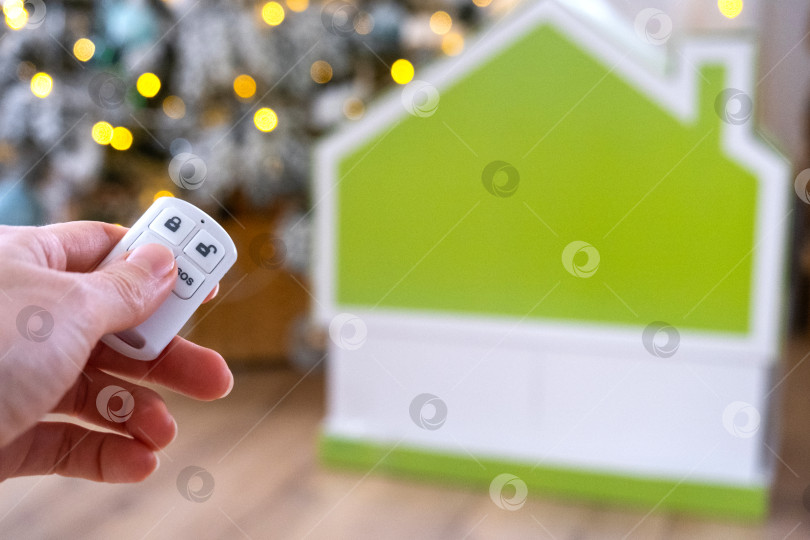 Скачать Брелок для системы безопасности в руке на фоне гирлянды рождественской елки и крошечного домика - подарок на Новый год, Рождество. Безопасность дома, сигнализация, сигнал SOS, защита от воров фотосток Ozero