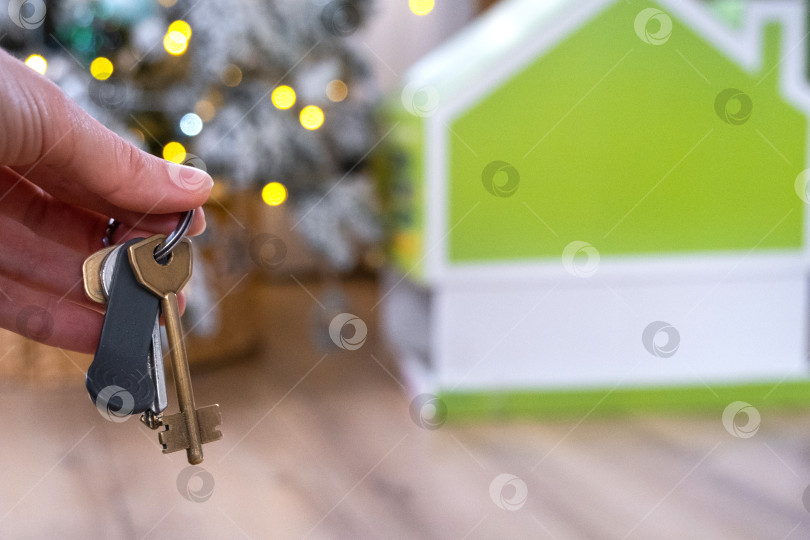 Скачать Ключи в руках на фоне рождественской елки с гирляндами и крошечного домика - подарок на Новый год, Рождество. Строительство, дизайн, проектирование, переезд, страхование, ипотека, аренда и покупка недвижимости. фотосток Ozero