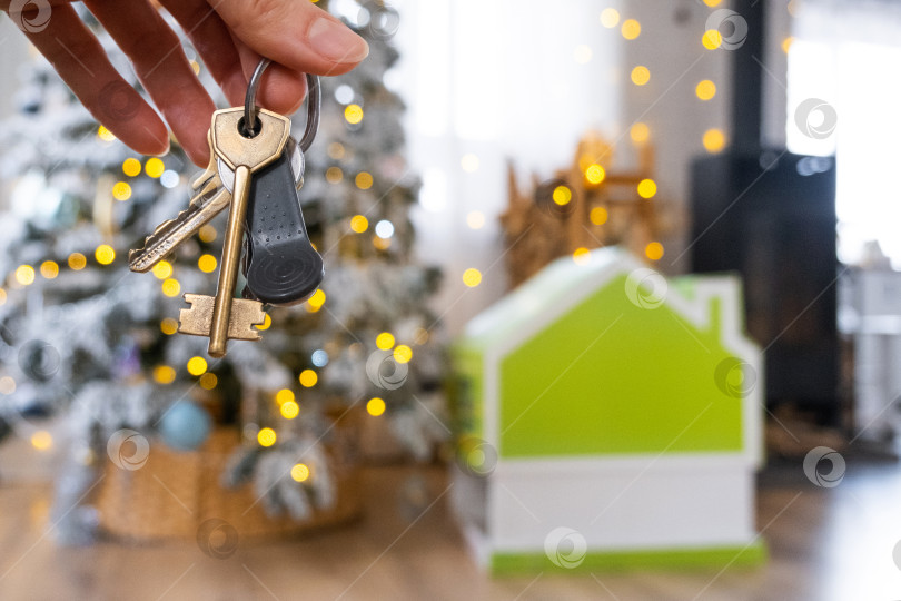 Скачать Ключи в руках на фоне рождественской елки с гирляндами и крошечного домика - подарок на Новый год, Рождество. Строительство, дизайн, проектирование, переезд, страхование, ипотека, аренда и покупка недвижимости. фотосток Ozero