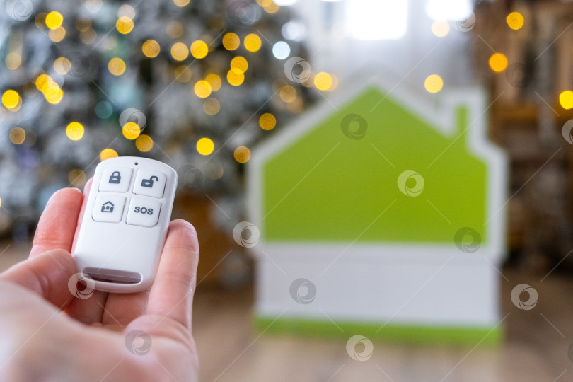 Скачать Брелок для системы безопасности в руке на фоне гирлянды рождественской елки и крошечного домика - подарок на Новый год, Рождество. Безопасность дома, сигнализация, сигнал SOS, защита от воров фотосток Ozero