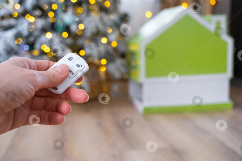 Скачать Брелок для системы безопасности в руке на фоне гирлянды рождественской елки и крошечного домика - подарок на Новый год, Рождество. Безопасность дома, сигнализация, сигнал SOS, защита от воров фотосток Ozero