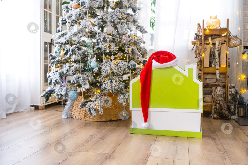 Скачать Декор рождественской елки в виде крошечного домика в шапке Санты в интерьере дома. Подарок на Новый год. Страхование, переезд в новый дом, ипотека, аренда и покупка недвижимости фотосток Ozero