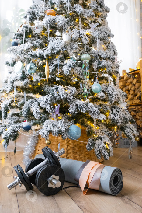 Скачать Спортивный инвентарь - гантели, коврик для фитнеса и ленты для фитнеса - лежат дома под рождественской елкой в качестве подарка на Рождество и Новый год фотосток Ozero
